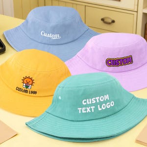May include: Four colorful bucket hats in blue, yellow, purple, and teal. The blue hat has "Custom" embroidered on it, the yellow hat has a sun logo, the purple hat has "Custom" in a bubble, and the teal hat says "Custom Text Logo".