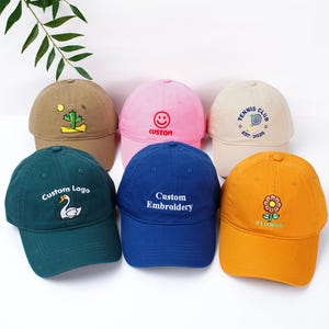 Könnte beinhalten: Sechs Baseballkappen in verschiedenen Farben: Beige, Rosa, Creme, Grün, Blau und Gelb. Jede Kappe hat eine individuelle Stickerei oder ein grafisches Design, darunter ein Kaktus, ein Smiley, ein Tennisclub-Logo, ein Schwan und eine Blume.