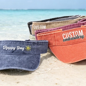 Könnte beinhalten: Eine Auswahl an Sonnenblenden in verschiedenen Farben, darunter Blau, Orange und Beige, liegt an einem Sandstrand. Auf den Sonnenblenden stehen die Wörter "Happy Day", "Custom" und "Custom embroidery". Im Hintergrund sind Meer und Himmel zu sehen.