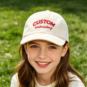 May include: A cream-colored baseball cap with the words "CUSTOM embroidery" embroidered in red. The cap is worn by a person with long brown hair and blue eyes, smiling in front of a green grassy background.