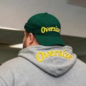 Op de afbeelding: Een donkergroene baseballcap met het woord "Oversize" in gele letters. De persoon draagt een grijze hoodie met het woord "Oversize" in gele letters op de capuchon.