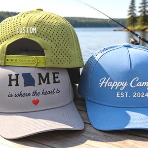 May include: A light green perforated baseball cap with a snapback closure and the word CUSTOM, a grey baseball cap with the text HOME is where the heart is and a blue heart, and a light blue baseball cap with the text Happy Camper EST. 2024.