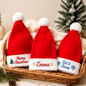 Może przedstawiać: Trzy czerwone czapki z dzianiny z białymi pomponami i białymi ściągaczami. Pierwsza czapka ma zielone drzewko i napis Merry Christmas. Druga czapka zawiera imię Emma ze złotymi płatkami śniegu. Trzecia czapka pokazuje niebieskie płatki śniegu i napis Let it Snow.