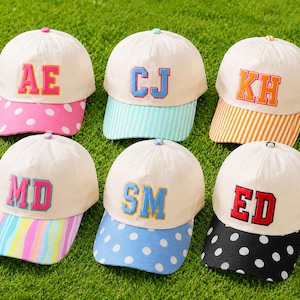 Könnte beinhalten: Sechs Baseballkappen mit cremeweißen Kronen und farbigen Schirmen. Die Kappen haben gestickte zweibuchstabige Monogramme in verschiedenen Farben. Die Schirme zeigen Muster wie weiße Punkte auf Pink, blaue Streifen, orange Streifen, mehrfarbige Wellen, weiße Punkte auf Blau und weiße Punkte auf Schwarz.