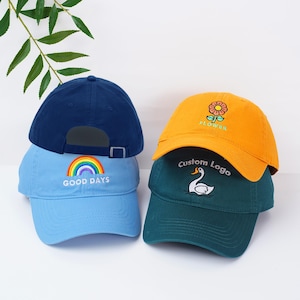 May include: A stack of four baseball caps in various colors. The top cap is navy blue, followed by a light blue cap with a rainbow and the words "GOOD DAYS". The next cap is orange with a flower design, and the bottom cap is teal with a swan and the words "Custom Logo".