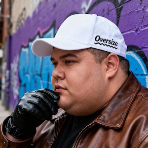 May include: A white baseball cap with the word "Oversize" in black script. The cap has a curved brim and a classic six-panel design. The hat is made of a durable fabric and is suitable for everyday wear.