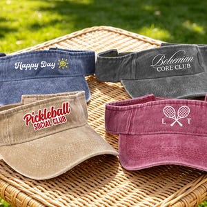 May include: Four visors in various colors are displayed on a wicker table. The visors are blue, gray, tan, and maroon. Each visor has text, including "Happy Day," "Bohemian Core Club," "Pickleball Social Club," and "Custom."