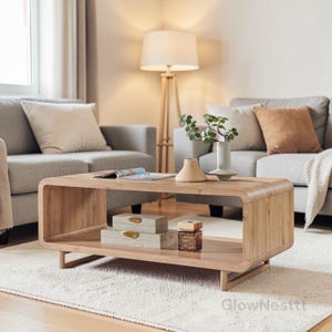 May include: A light-colored wooden coffee table with a rounded design. The table has a lower shelf with two decorative boxes and a perfume bottle. The table is in a living room setting with a gray sofa and a floor lamp.