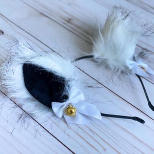 May include: White furry cat ears headband with black velvet ears and white bows with a gold bell.