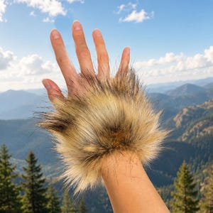 May include: A wrist cuff made of long, shaggy faux fur in shades of tan, beige, and dark brown. The accessory features a dense, textured pile that encircles the wrist, providing a thick layer of synthetic fibers with varied color tones.