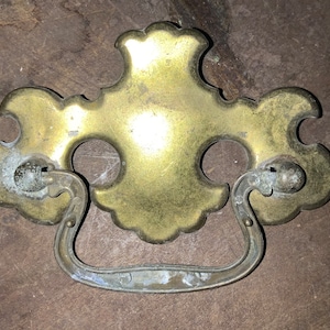 May include: A vintage brass cabinet pull with a decorative, ornate design. The pull has a loop handle and is in a worn, antique finish.