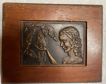 May include: A wooden box with a raised, bronze-colored plaque depicting a woman with a braid and a parrot. The plaque is set against a background of stylized banana leaves.