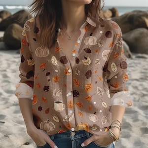 May include: A button-down shirt with a brown background and a fall-themed pattern. The pattern includes pumpkins, leaves, and coffee mugs in shades of orange, brown, and white. The shirt has long sleeves and a collared neckline.