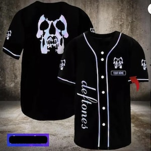 May include: Black baseball jersey with white trim and buttons. The jersey features a skull design on the front and back, with the word "Deftones" in white script. A small patch with "YOUR NAME" is also visible.