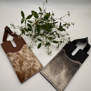 May include: Two rectangular luggage tags featuring a top section of smooth leather and a bottom section of textured animal hide. One tag is brown with a tan and white hide, while the other is black with a grey and white hide, each with a snap closure.