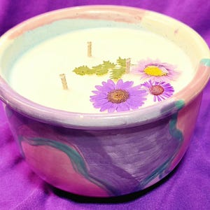 May include: A handmade candle in a ceramic bowl with a painted design. The candle wax is white, with three wicks and decorated with dried flowers. The bowl is painted with pink, purple, and teal colors.