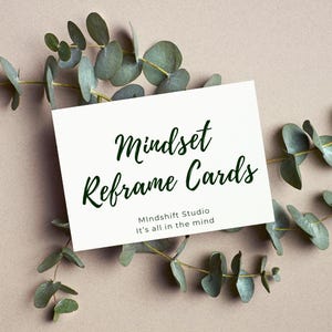 May include: A white card with the text "Mindset Reframe Cards" in dark green script, with the text "Mindshift Studio" and "It's all in the mind" below. The card is surrounded by green eucalyptus leaves on a neutral background.
