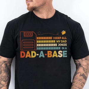 Op de afbeelding: Zwart t-shirt met een retro ontwerp met een floppy disk-afbeelding en de tekst "I keep all my dad jokes in a DAD-A-BASE." Het ontwerp gebruikt oranje, geel, blauw en groen.