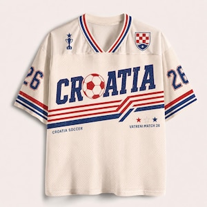 K&ouml;nnte beinhalten: Ein cremefarbenes Mesh-Trikot mit dem Schriftzug CROATIA, bei dem der Buchstabe O als Fu&szlig;ball dargestellt ist. Das Shirt zeigt rote und blaue Streifen, die Nummer 26 auf den &Auml;rmeln, ein Pokalsymbol, das kroatische Wappen und den Text VATRENI MATCH 26.