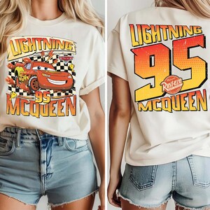 May include: Cream-coloured t-shirt featuring a graphic of Lightning McQueen from the Cars film. The front displays the character with the text "Lightning 95 McQueen." The back of the shirt has "Lightning 95 McQueen" in a gradient orange and yellow design.