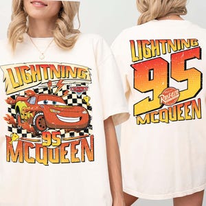 May include: A cream-coloured t-shirt with a graphic of Lightning McQueen from the film Cars. The front design features "Lightning McQueen 95" with a chequered flag background. The back of the shirt displays "Lightning 95 McQueen" in a gradient orange and yellow.
