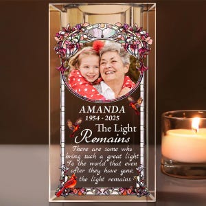 May include: A clear glass candle holder with a photo of two people and the text "AMANDA 1954-2025 The Light Remains". The holder has a stained glass design with red and purple accents and a small candle is lit to the right.
