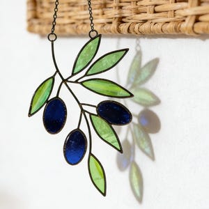 Puede incluir: Una rama de olivo de vidriera con hojas verdes y aceitunas azules, colgada de una cadena negra. La pieza decorativa está colocada contra una pared blanca y una cesta de mimbre.