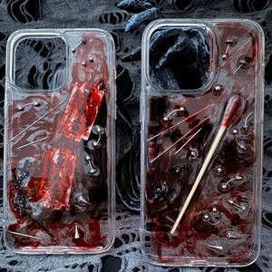 Peut inclure: Deux coques de téléphone transparentes avec une substance rouge foncé semblable à du sang à l'intérieur. L'une contient des lames de rasoir, l'autre une allumette. Les deux coques ont des clous et des piercings argentés.