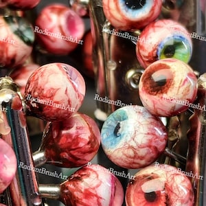 May include: A collection of handmade eyeball beads in shades of red, white, and blue. The beads feature detailed vein patterns and are attached to metal findings. The text "RadiantBrushArt" is visible on some of the beads. The beads are approximately 1-2 cm in diameter.