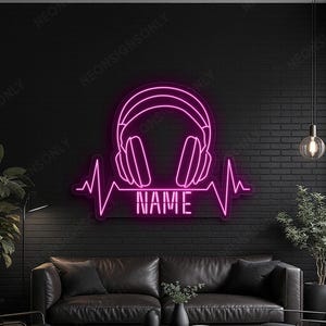 Puede incluir: Un letrero de neón rosa que representa unos auriculares sobre una línea de latidos del corazón, con la palabra "NAME" en blanco. El letrero está montado en una pared de ladrillo negro. La habitación está amueblada con un sofá de cuero negro, sillones y una mesa de centro oscura.