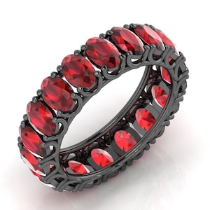 May include: A black eternity ring featuring oval-cut, vibrant red gemstones. The ring's dark metal setting is intricately designed, showcasing the gemstones in a continuous loop. The ring is displayed on a white background.