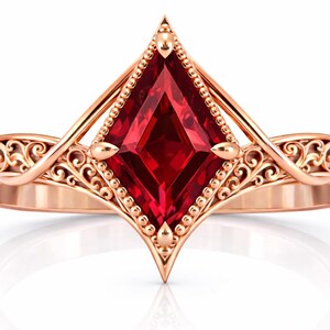 May include: Rose gold ring featuring a large, vibrant red gemstone in a kite shape. The ring has intricate filigree detailing along the band and around the gemstone, with small beaded accents. The design is elegant and ornate, suitable for special occasions.