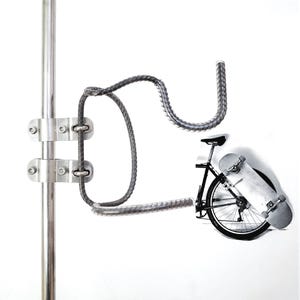 Puede incluir: Un soporte de pared de metal con una barra vertical y un brazo curvo de varilla corrugada para sujetar un monopatín. El soporte se fija con dos abrazaderas de metal plateado. Una imagen pequeña muestra una bicicleta con un monopatín.