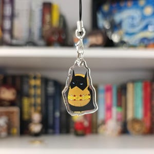 以下が含まれることがあります： バットマンをテーマにしたキーホルダーチャーム。チャームは、バットマンのロゴの黄色と黒のデザインが特徴です。チャームは、本や装飾品でいっぱいの本棚の前に吊り下げられています。