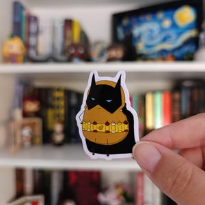以下が含まれることがあります： バットマンにインスパイアされたコスチュームを着た漫画のキャラクターのステッカー。キャラクターは黒いマスク、青い目、黄色のユーティリティベルトをしています。ステッカーは本棚の前に保持されています。