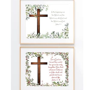 May include: Two framed wall art prints featuring a dark wood cross surrounded by green eucalyptus leaves. The top print displays the text In the beginning was the Word and the Word was with God and the Word was God John 1:1. The bottom print displays For God so loved the world he gave his only begotten Son, that whoever believes in Him shall not perish but have eternal life John 3:16.
