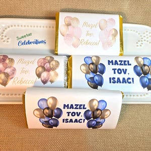 May include: Chocolate bar wrappers with balloon designs and the text "Mazel Tov, Rebecca!" and "Mazel Tov, Isaac!" in blue, gold, and pink. The wrappers are on a white decorative tray.