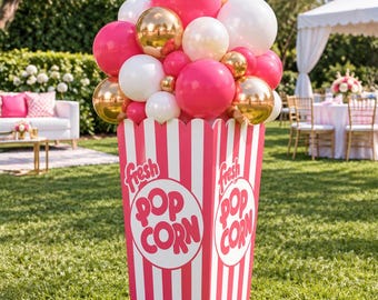 以下が含まれることがあります： ピンクと白の縦縞模様で「fresh POP CORN」という文字がピンク色で書かれた、大きなポップコーン容器の装飾。上部には、ホットピンク、白、メタリックゴールドの丸い風船の束があふれるように飾られています。