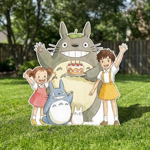 Peut inclure: Une grande découpe plate représentant un Totoro gris tenant un gâteau d'anniversaire avec des bougies, accompagné d'un plus petit Totoro bleu, d'un minuscule Totoro blanc et de deux enfants aux cheveux bruns, l'un en robe rose et l'autre en jupe jaune.