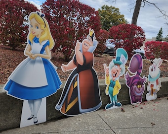 May include: Cardboard cutouts of Alice in Wonderland characters, including Alice, the Queen of Hearts, the Mad Hatter, the Cheshire Cat, and the White Rabbit. The characters are brightly colored and stand on a concrete surface.