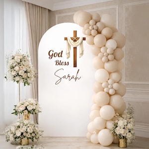 May include: A white arched backdrop with a wooden cross and the words "God Bless Sarah" in brown script. A garland of beige balloons and floral arrangements frame the backdrop, creating a celebratory atmosphere.