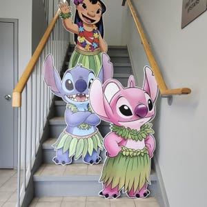 May include: Cardboard cutouts of Lilo, Stitch, and Angel from the Disney movie Lilo & Stitch. Lilo is waving, wearing a red top, flower lei, and green skirt. Stitch and Angel are wearing leis and grass skirts. The cutouts are on a staircase.