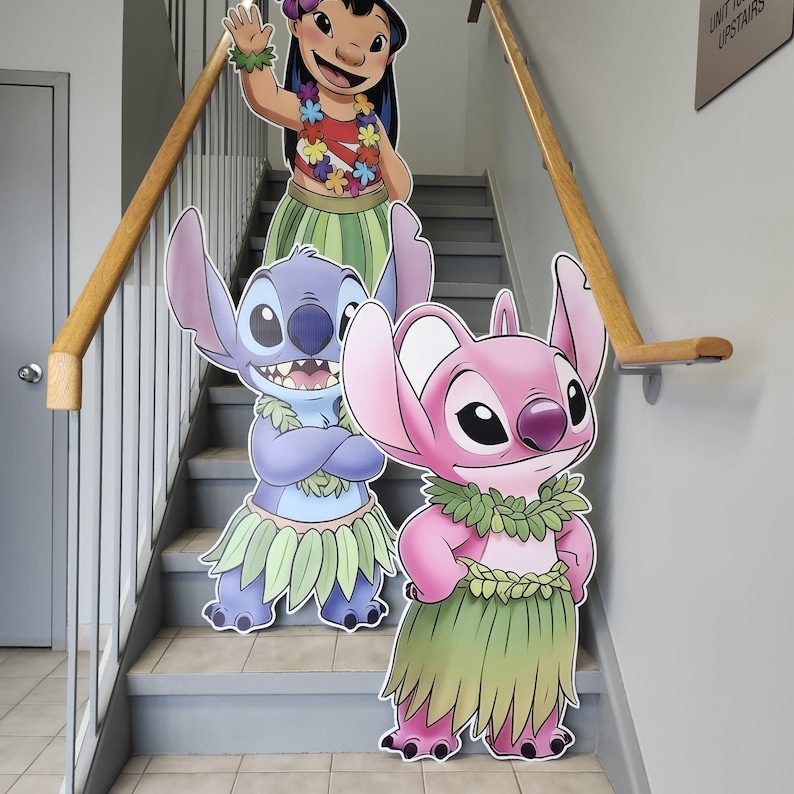 May include: Cardboard cutouts of Lilo, Stitch, and Angel from the Disney movie Lilo & Stitch. Lilo is waving, wearing a red top, flower lei, and green skirt. Stitch and Angel are wearing leis and grass skirts. The cutouts are on a staircase.