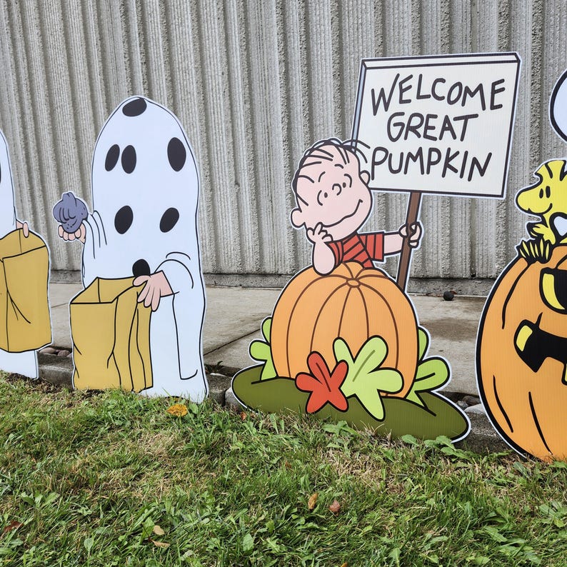 May include: A set of Peanuts-themed yard signs featuring a character in a ghost costume with black spots holding a tan bag, and a character leaning on a large orange pumpkin while holding a sign that reads WELCOME GREAT PUMPKIN with a yellow bird.