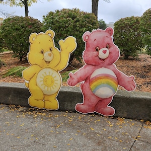Peut inclure: Deux découpes de Care Bears. L'une est jaune avec un soleil souriant sur le ventre, levant sa patte droite. L'autre est rose avec un arc-en-ciel sur le ventre, les bras ouverts. Les deux ont des visages souriants.