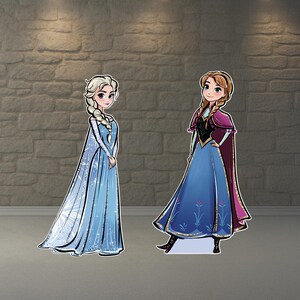 May include: Two cardboard cutouts of animated characters, Elsa and Anna, from the movie Frozen. Elsa is in a blue dress with a long cape, and Anna is in a blue dress with a purple cape. Both are standing on a gray surface.