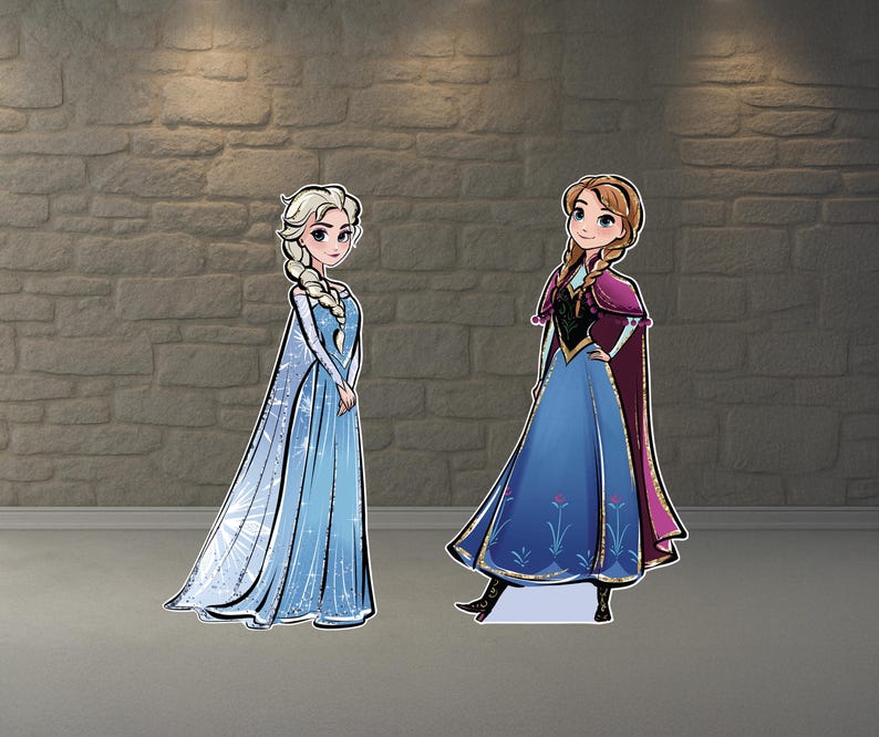 May include: Two cardboard cutouts of animated characters, Elsa and Anna, from the movie Frozen. Elsa is in a blue dress with a long cape, and Anna is in a blue dress with a purple cape. Both are standing on a gray surface.