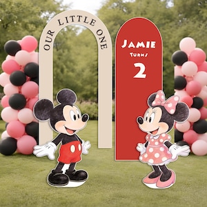 May include: A birthday party scene featuring Mickey and Minnie Mouse cutouts, a red sign reading "Jamie Turns 2", and an arch with the words "Our Little One". Pink, black, and white balloons are arranged in the background. The setting is a grassy lawn.