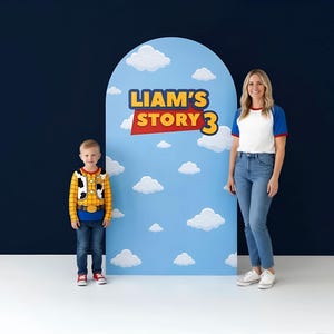 May include: An arched backdrop featuring a light blue sky with white clouds and the text LIAM'S STORY 3 in yellow and red lettering. The backdrop is positioned between a child wearing a yellow plaid shirt and an adult in a white and blue shirt.