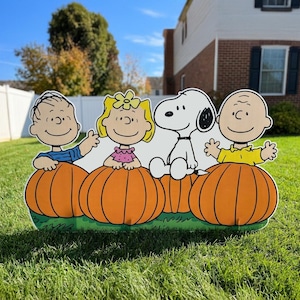 Puede incluir: Un letrero decorativo para jardín con los personajes de dibujos animados Linus, Sally, Snoopy y Charlie Brown sentados detrás de cuatro calabazas naranjas grandes. Los personajes tienen contornos negros sobre un fondo blanco.